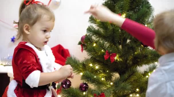 宝宝和妈妈在圣诞树上挂着红球玩具。快乐童年的概念。孩子和母亲用圣诞球装饰圣诞树。小孩和父母正在圣诞树旁玩耍. — 图库视频影像