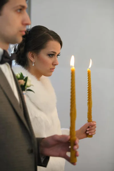 Noiva e noivo em pé na cerimônia de casamento. Casal de casamento elegante feliz segurando velas com luz sob coroas douradas durante o matrimônio sagrado na igreja . — Fotografia de Stock