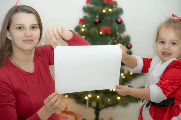 Mama i córka ubrane na czerwono trzymać biały arkusz papieru na tle choinki — Zdjęcie stockowe