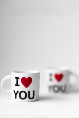 Kırmızı kalpli iki beyaz bardak ve bir yazı. Beyaz arka planda seni seviyorum.