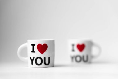 Kırmızı kalpli iki beyaz bardak ve bir yazı. Beyaz arka planda seni seviyorum.