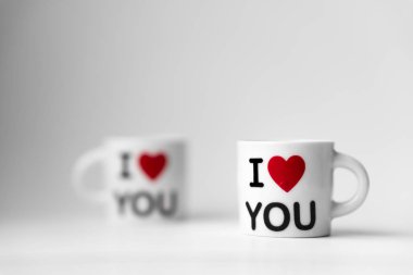 Kırmızı kalpli iki beyaz bardak ve bir yazı. Beyaz arka planda seni seviyorum.