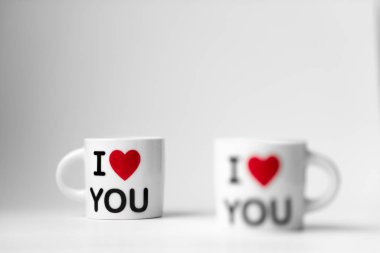 Kırmızı kalpli iki beyaz bardak ve bir yazı. Beyaz arka planda seni seviyorum.
