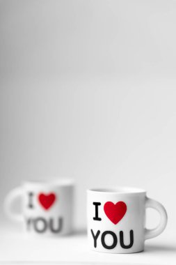 Kırmızı kalpli iki beyaz bardak ve bir yazı. Beyaz arka planda seni seviyorum.
