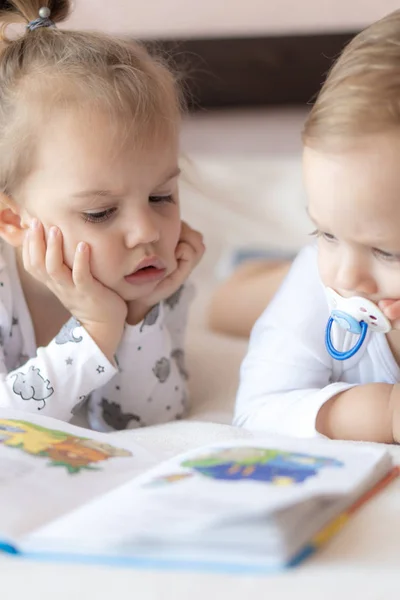 Niños encantadores - hermano y hermana, leyendo un libro, en la cama. Primer plano de los niños en la cama leyendo un libro. Un niño y una niña de blanco están jugando en la cama. Bebés de blanco en la cama. Lindos niños . — Foto de Stock