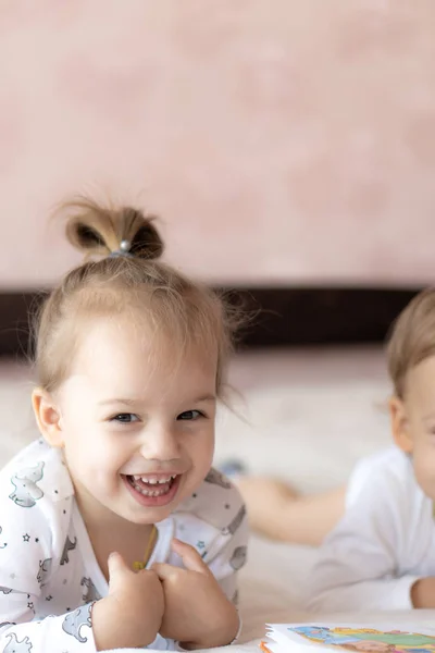 Niños encantadores - hermano y hermana, leyendo un libro, en la cama. Primer plano de los niños en la cama leyendo un libro. Un niño y una niña de blanco están jugando en la cama. Bebés de blanco en la cama. Lindos niños . — Foto de Stock