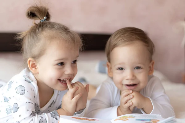 Krásné děti - bratr a sestra, čtení knihy, na posteli. Zblízka si děti v posteli čtou knížku. Chlapec a dívka v bílém si hrají na posteli. Děti v bílém na posteli. Roztomilé děti. — Stock fotografie