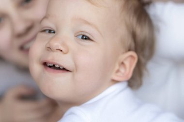 Croon-plan toddler in white looks up and smiles in front of mother