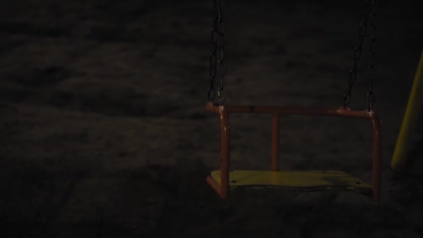 Swing en el patio de recreo por la noche, nadie en el parque. Paseos en primer plano en la oscuridad. El columpio se balancea lentamente. Zona de juegos abandonada por la noche. Paisaje nocturno sombrío en el parque . — Vídeos de Stock