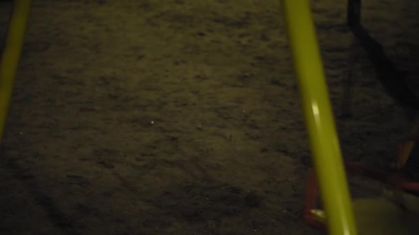 Swing on the playground at night, no one in the park. Close-up rides in the dark. The swing sways slowly. Deserted playground at night. Gloomy night scenery in the park. — Stock Video