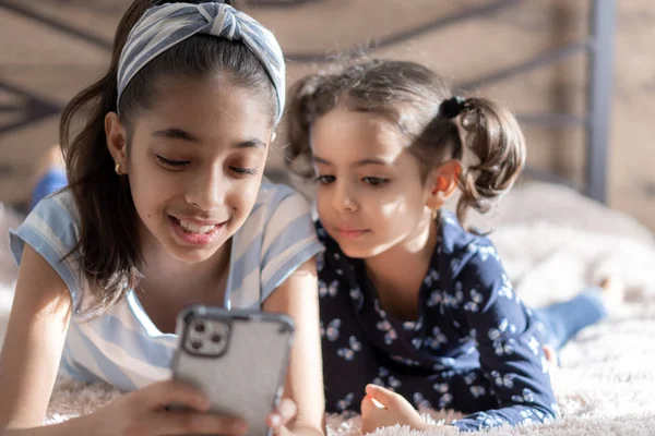 Dos niñas de piel oscura, hermanas, están acostadas en la cama y mirando un teléfono inteligente contra el fondo del sol desde la ventana. Chicas persas en la cama con un teléfono. Niños de Oriente Medio . — Foto de Stock