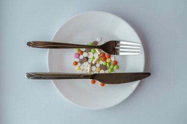 Beyaz tabakta haplar. Bıçak ve çatalla yapılan jest tabakta haplarla birlikte mükemmeldir. Tabakta çatal bıçakla yemek kılığına girmiş bir yığın renkli tablet..