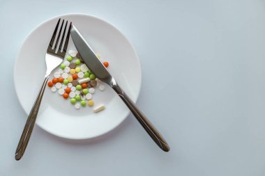 Beyaz tabakta haplar. Bıçak ve çatalla duraklat, tabakta haplarla birlikte. Beyaz arka planda ilaçlar. Tabakta çatal bıçakla yemek kılığına girmiş bir yığın renkli tablet..