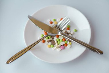 Beyaz tabakta haplar. Bıçak ve çatalla yapılan jest, bir tabak hapın üzerindeki yemeği bitirdi. İlaçlar beyaz tabakta. Tabakta çatal bıçakla yemek kılığına girmiş bir yığın renkli tablet..