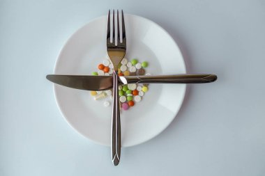 Beyaz tabakta haplar. Bıçak ve çatalla tabakta haplarla yapılan jestler. Beyaz arka planda ilaçlar. Tabakta çatal bıçakla yemek kılığına girmiş bir yığın renkli tablet..