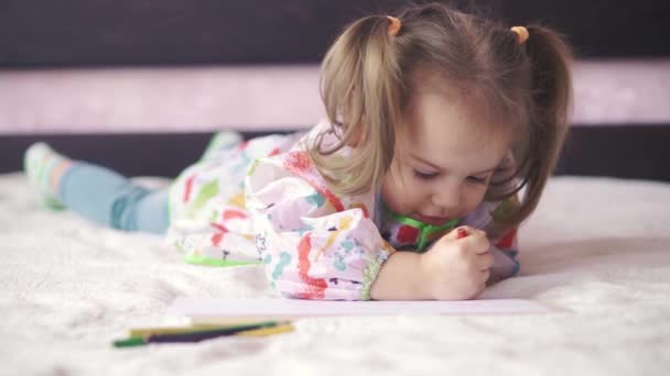 Primo piano di una bambina in vestaglia seduta su un divano e che si morde le labbra. Bambino carino con due trecce guarda attentamente la fotocamera . — Video Stock