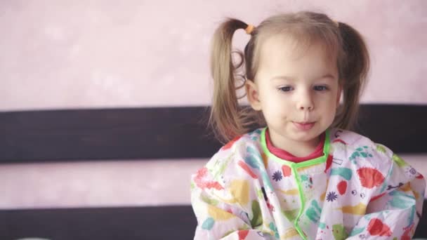 Close-up shot of a little girl in a dressing gown sitting on a sofa and biting her lips. Cute baby with two pigtails looks carefully at the camera. — Stock Video