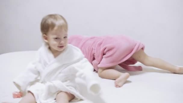 Δύο παιδιά με μπουρνούζια σε λευκό φόντο με βρεγμένα μαλλιά μετά το μπάνιο. Αγόρι και κορίτσι μετά το ντους σε λευκό κρεβάτι. Τα αδέρφια με τα μπουρνούζια παίζουν πριν κοιμηθούν. Παιδιά ιρλανδικά δίδυμα σε ένα — Αρχείο Βίντεο