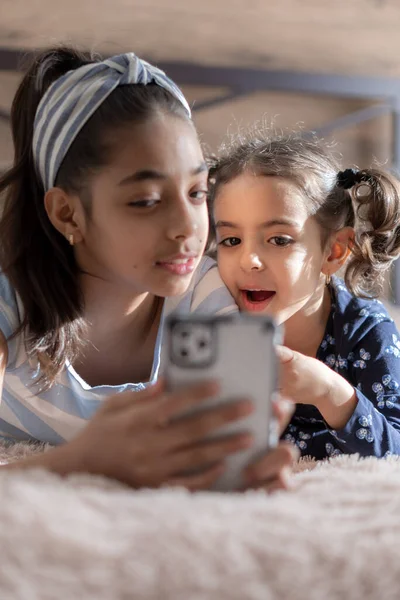 Δύο σκουρόχρωμες νεαρές κοπέλες, αδελφές, ξαπλώνουν στο κρεβάτι και κοιτάζουν ένα smartphone στο φόντο του ήλιου από το παράθυρο. Περσίδες στο κρεβάτι με τηλέφωνο. Μικρά παιδιά της Μέσης Ανατολής. — Φωτογραφία Αρχείου
