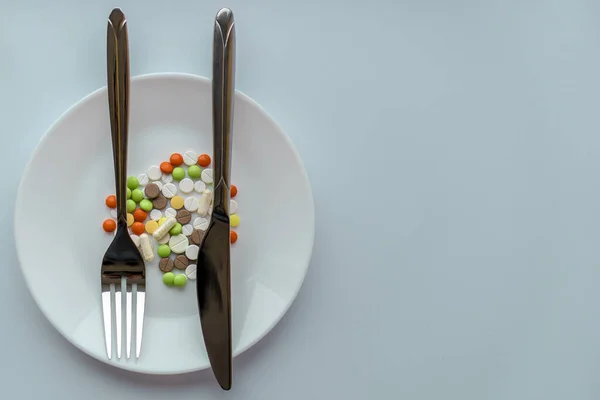 Pills on a white plate. Gesture with a knife and fork, bring the plaintive book, on a plate with pills. A pile of multi-colored tablets disguised as food on a plate with cutlery.