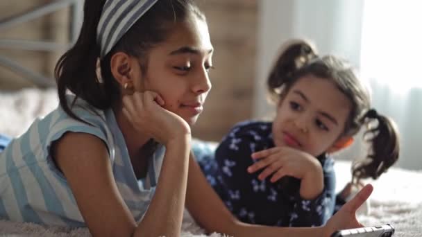 Zwei junge Mädchen aus dem Nahen Osten auf dem Bett mit einem Smartphone. Zwei dunkelhäutige Schwestern mit einer Tablettengrimasse und zeigen negative Emotionen. Zwei persische Mädchen spielen mit einem Smartphone auf dem Bett. — Stockvideo