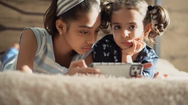 Deux jeunes filles du Moyen-Orient sur le lit avec un smartphone. Deux soeurs sur le lit s'embrassent. Deux sœurs noires se blottissent dans un lit lumineux. Deux filles persanes jouent avec un smartphone sur le lit . — Video