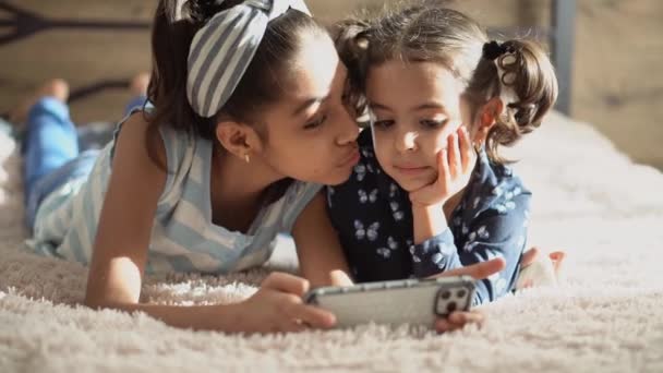 Zwei junge Mädchen aus dem Nahen Osten auf dem Bett mit einem Smartphone. Zwei finstere Schwestern auf dem Bett küssen sich. Das Mädchen versucht, ihre kleine Schwester zu küssen, und sie wendet sich ab. Zwei persische Mädchen spielen mit einem — Stockvideo