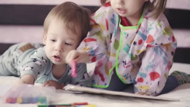 Zwei süße Kinder malen mit Kreonen, die auf dem Bett liegen. Mädchen und Jungen spielen auf dem Bett mit Bleistiften. Geschwister-Zwillinge zeichnen im Liegen mit Bleistiften. — Stockvideo