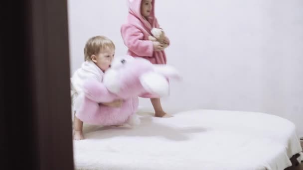 Two kids in bathrobes on a white background with wet hair after taking a bath. Boy and girl after shower on a white bed. Brother and sister in bathrobes play before bedtime. Children Irish twins on a — Stock Video