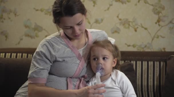 Mała dziewczynka robi inhalację za pomocą inhalatora sprężarki siedząc na kanapie. Dziecko w miejscu z ciężarną matką robi inhalację. Mama robi inhalację swojej córeczce. — Wideo stockowe