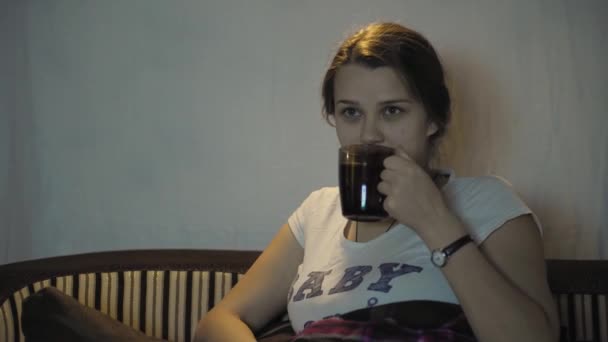 Linda chica embarazada viendo la televisión en una habitación oscura. Una mujer embarazada con una camiseta blanca con la inscripción en el pecho "bebé a bordo" está viendo la televisión. La chica mira la televisión y come . — Vídeos de Stock