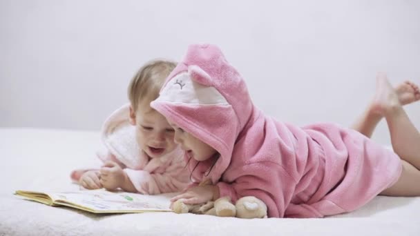 Two kids in bathrobes on a white background with wet hair after taking a bath. Boy and girl after shower on a white bed. Brother and sister in bathrobes play before bedtime. Children Irish twins on a — Stock Video