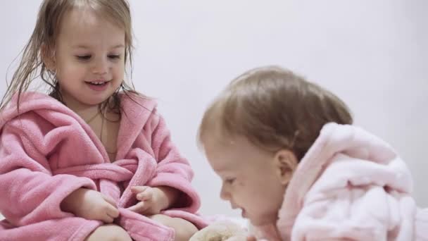 Two kids in bathrobes on a white background with wet hair after taking a bath. Boy and girl after shower on a white bed. Brother and sister in bathrobes play before bedtime. Children Irish twins on a — Stockvideo