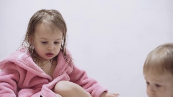 Two kids in bathrobes on a white background with wet hair after taking a bath. Boy and girl after shower on a white bed. Brother and sister in bathrobes play before bedtime. Children Irish twins on a — Stockvideo