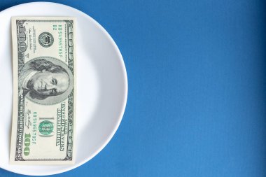 İlaçlar paraya mal olur. Uyuşturucu ve dolarlar. Beyaz plaka, çok renkli haplar, kapsüller ve klasik mavi arka planda 100 dolarlık banknot. Haplar ve para mavi arka planda beyaz bir tabakta.