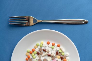 Beyaz tabakta haplar. Tabakta çatal ve haplarla yapılan jestler. Klasik mavi arka planda ilaçlar. Tabakta çatal bıçakla yemek kılığına girmiş bir yığın renkli tablet..