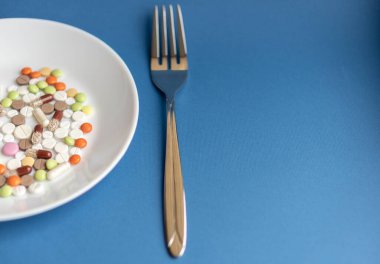 Beyaz tabakta haplar. Tabakta çatal ve haplarla yapılan jestler. Klasik mavi arka planda ilaçlar. Tabakta çatal bıçakla yemek kılığına girmiş bir yığın renkli tablet..