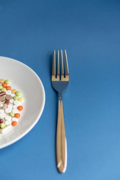 Pills on a white plate. Gestures with a fork on a plate with pills. Medicines on a classic blue background. A pile of multi-colored tablets disguised as food on a plate with cutlery.
