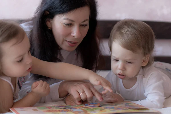 Babička s vnukem a vnučkou v posteli čtou knížku. Babička s vnoučaty v posteli během dálkového studia. Babička si hraje se svými vnoučaty v posteli během karantény. — Stock fotografie