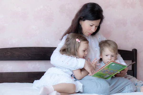 Η γιαγιά με τον εγγονό και την εγγονή στο κρεβάτι διαβάζουν ένα βιβλίο. Γιαγιά με εγγόνια στο κρεβάτι κατά τη διάρκεια της εξ αποστάσεως εκπαίδευσης. Η γιαγιά παίζει με τα εγγόνια της στο κρεβάτι κατά τη διάρκεια της απομόνωσης. — Φωτογραφία Αρχείου