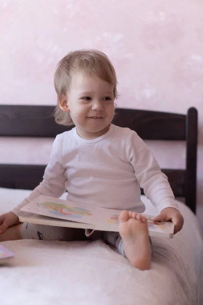 Um menino loiro está sentado na cama e sorrindo, segurando um livro em suas mãos. Um menino se senta em uma cama com um livro em suas mãos durante a quarentena . — Fotografia de Stock