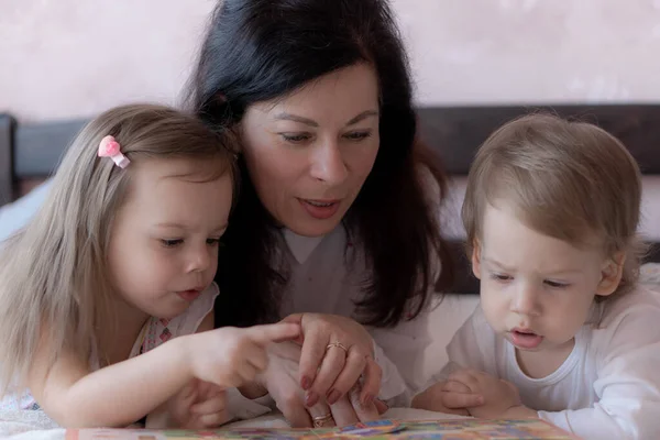 Grand-mère avec petit-fils et petite-fille au lit lisant un livre. Grand-mère avec petits-enfants au lit pendant l'enseignement à distance. Grand-mère joue avec ses petits-enfants au lit pendant la quarantaine . — Photo