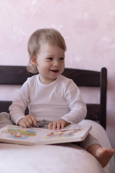 Um menino loiro está sentado na cama e sorrindo, segurando um livro em suas mãos. Um menino se senta em uma cama com um livro em suas mãos durante a quarentena . — Fotografia de Stock