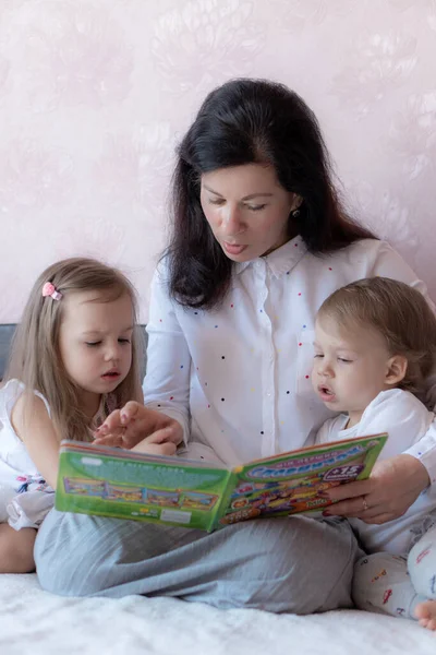 Η γιαγιά με τον εγγονό και την εγγονή στο κρεβάτι διαβάζουν ένα βιβλίο. Γιαγιά με εγγόνια στο κρεβάτι κατά τη διάρκεια της εξ αποστάσεως εκπαίδευσης. Η γιαγιά παίζει με τα εγγόνια της στο κρεβάτι κατά τη διάρκεια της απομόνωσης. — Φωτογραφία Αρχείου