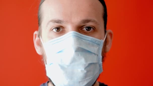 Coronavirus, epidemic, pandemic, quarantine, self-isolation, security, protection, medicine concept - long-haired bearded thin young man in medical protective mask on plain red background close-up. — Stock Video