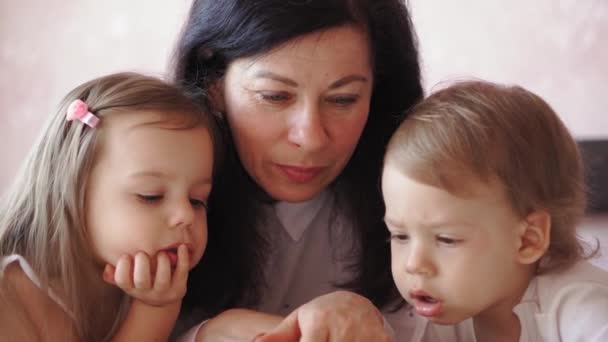 Familia, infancia, maternidad, crianza, autoaislamiento, concepto de aprendizaje a distancia - Abuela y nietos niña niño lee libro en la cama. Mujer mayor, niños. Niñera mayor, los niños juegan en casa . — Vídeos de Stock