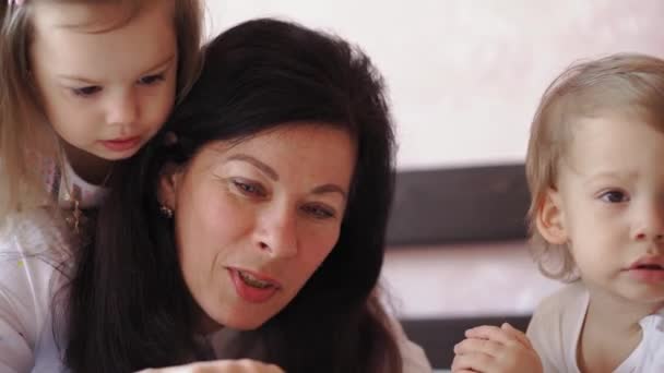 Familia, infancia, maternidad, crianza, autoaislamiento, concepto de aprendizaje a distancia - Abuela y nietos niña niño lee libro en la cama. Mujer mayor, niños. Niñera mayor, los niños juegan en casa . — Vídeos de Stock