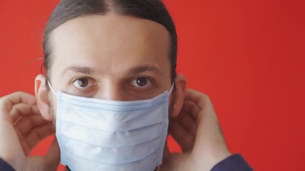 Covid-19, epidemic, pandemic, quarantine, self-isolation, protection, medicine concept - portrait of long-hair-bearded man wearing blue protective medical mask on plain red background close-up, copy spase. — 图库视频影像