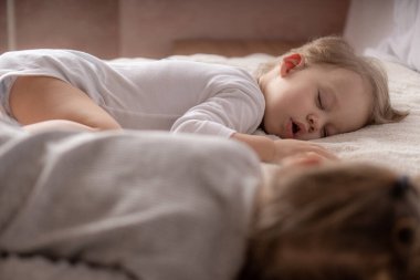 Çocukluk, uyku, rahatlama, aile, yaşam tarzı konsepti - 2 ve 3 yaşlarındaki iki genç, beyaz ve bej giysiler içinde öğle yemeğinde el ele tutuşarak bej ve beyaz bir yatakta uyurlar..