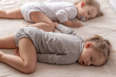 Çocukluk, uyku, rahatlama, aile, yaşam tarzı konsepti - 2 ve 3 yaşlarındaki iki genç, beyaz ve bej giysiler içinde öğle yemeğinde el ele tutuşarak bej ve beyaz bir yatakta uyurlar..